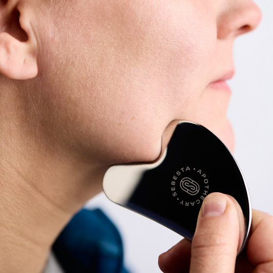 Stainless steel Gua Sha with Sebesta Apothecary logo being used on woman's chin.