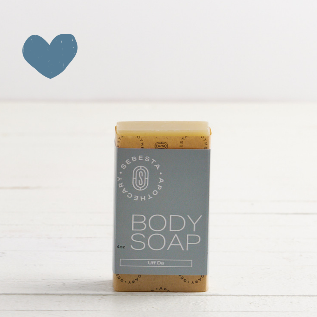 Sebesta Apothecary Body Soap Bar, with blue wrap on white table with white background. Sent called Uff Da. Blue heart icon top left