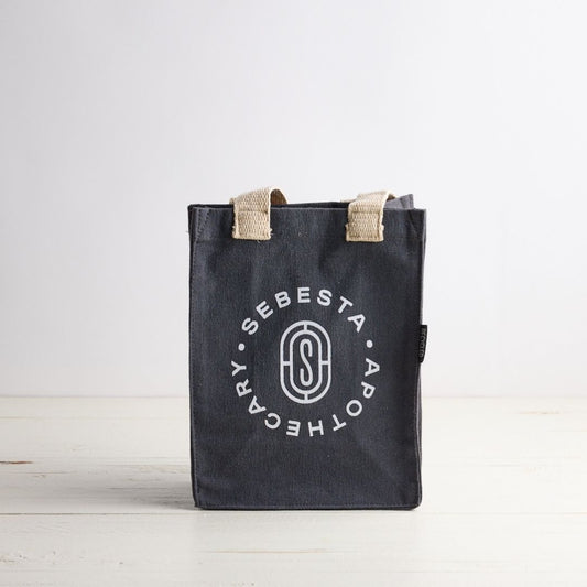 Blue gray tote bag with white Sebesta Apothecary logo, cream fabric handles. Set on white wood table and white background.