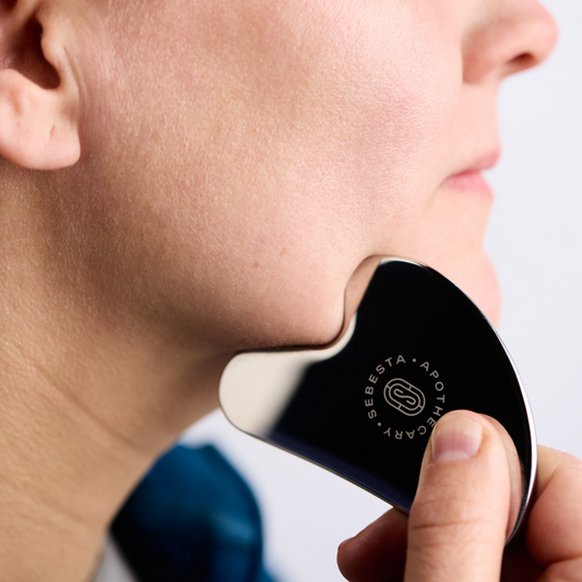 Stainless steel Gua Sha with Sebesta Apothecary logo being used on woman's chin. 