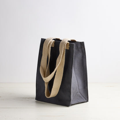 Blue gray tote bag with white Sebesta Apothecary logo, cream fabric handles. Set on white wood table and white background.
