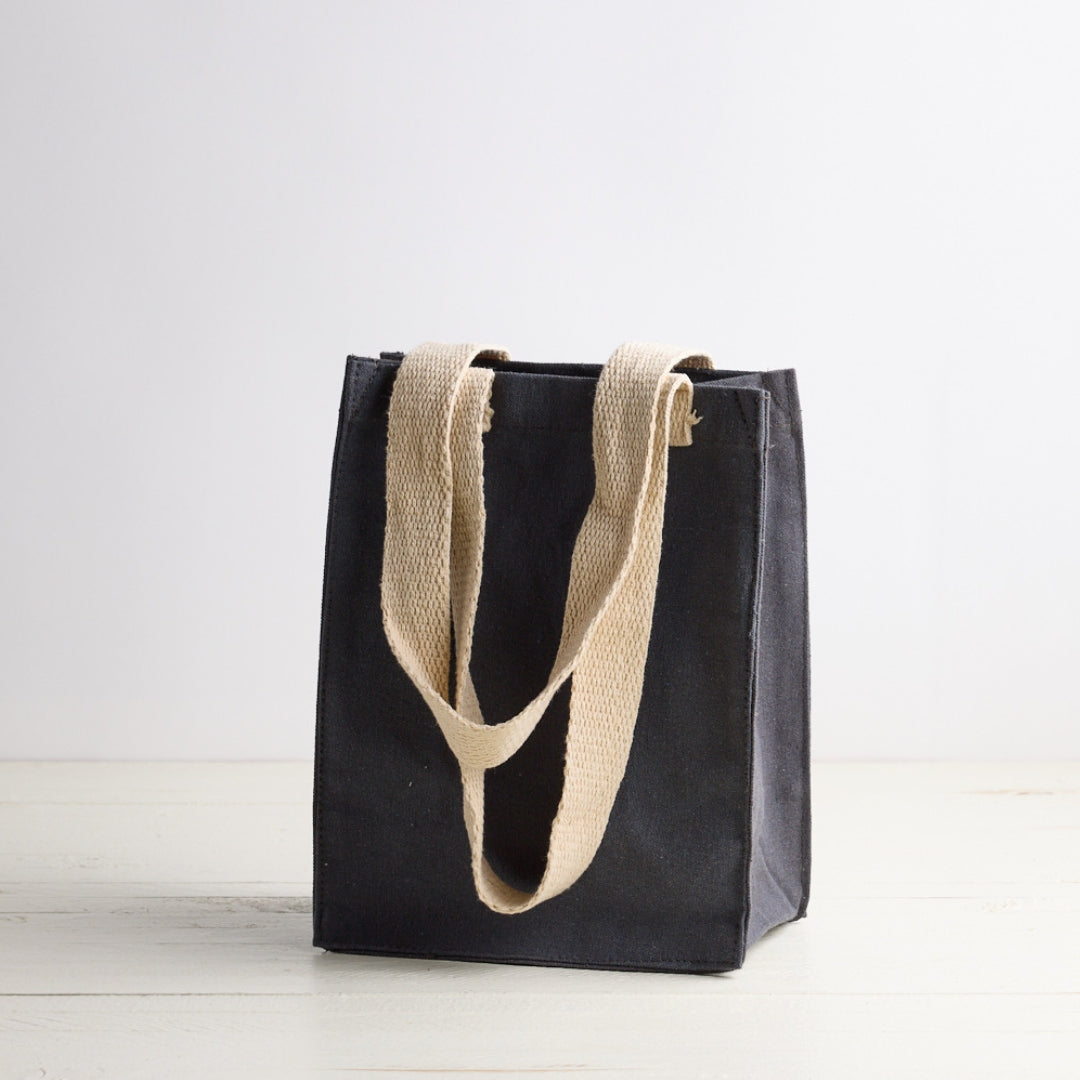 Blue gray tote bag with white Sebesta Apothecary logo, cream fabric handles. Set on white wood table and white background.