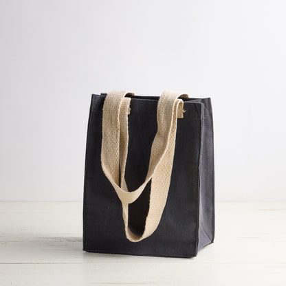 Blue gray tote bag with white Sebesta Apothecary logo, cream fabric handles. Set on white wood table and white background.