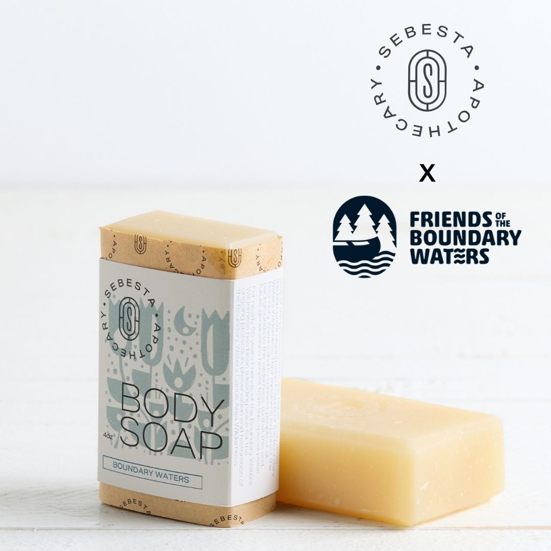 Sebesta Apothecary Soap bars - one standing with label facing forward the other unlabeled on table. White background and table. Logos top right for both Sebesta Apothecary and the Friends of the Boundary Waters