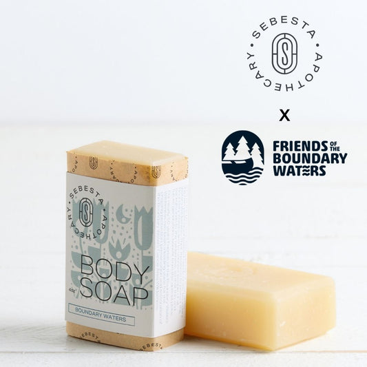 Sebesta Apothecary Soap bars - one standing with label facing forward the other unlabeled on table. White background and table. Logos top right for both Sebesta Apothecary and the Friends of the Boundary Waters