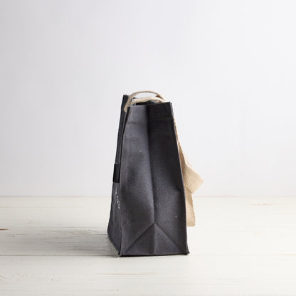 Blue gray tote bag with white Sebesta Apothecary logo, cream fabric handles. Set on white wood table and white background.