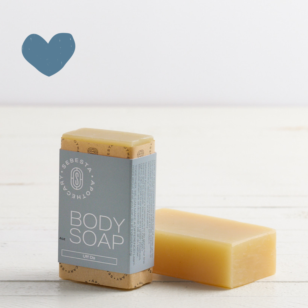 Sebesta Apothecary Body Soap Bar, with blue wrap another next to it laying down no label. On white table with white background. Sent called Uff Da. Blue heart icon top left