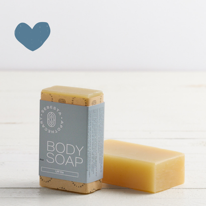 Sebesta Apothecary Body Soap Bar, with blue wrap another next to it laying down no label. On white table with white background. Sent called Uff Da. Blue heart icon top left