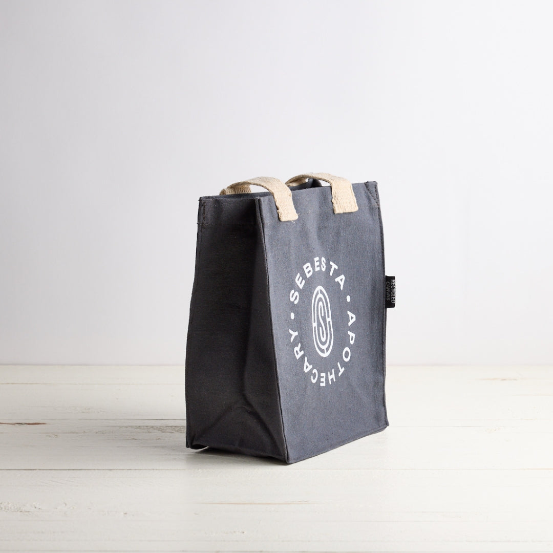Gray tote bag with beige handles on a light wooden surface. Sebesta Apothecary on front