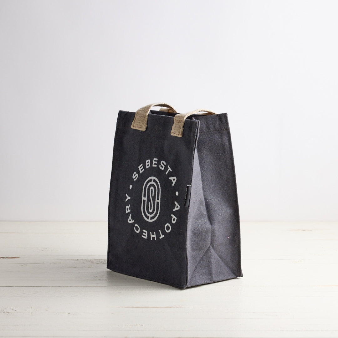 Blue gray tote bag with white Sebesta Apothecary logo, cream fabric handles. Set on white wood table and white background.