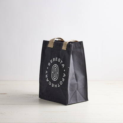 Blue gray tote bag with white Sebesta Apothecary logo, cream fabric handles. Set on white wood table and white background.