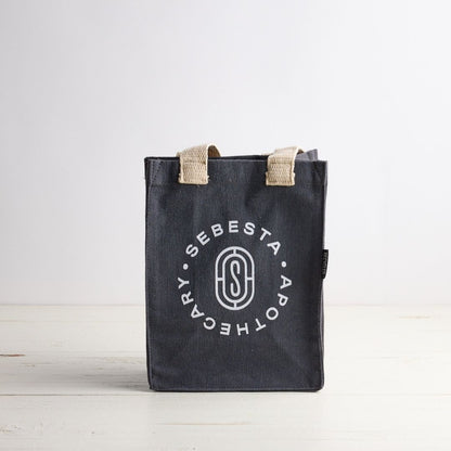 Blue gray tote bag with white Sebesta Apothecary logo, cream fabric handles. Set on white wood table and white background.