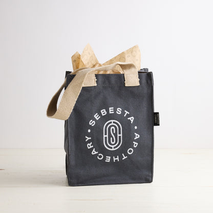Black tote bag with 'Sebesta Apothecary' logo on a white background